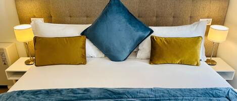 A bed with a blue duvet, two white pillows, and two gold pillows. There are two bedside lamps with white shades.