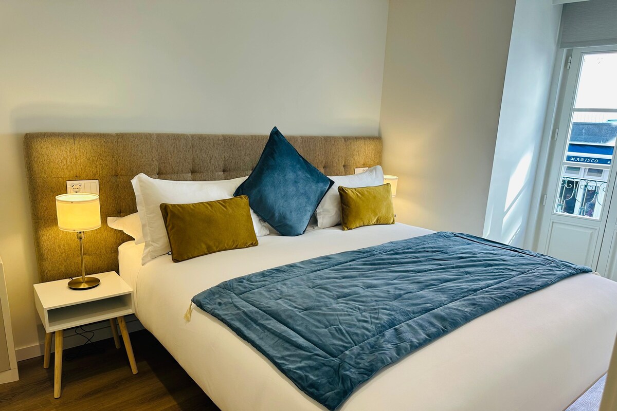 A hotel room with a bed, a nightstand with a lamp, and a window with a view.