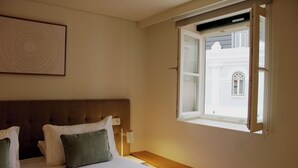 A hotel room with a bed, pillows, a nightstand, a lamp, and a window with a view of a building. - Rossio Garden Suites (Lisbon)