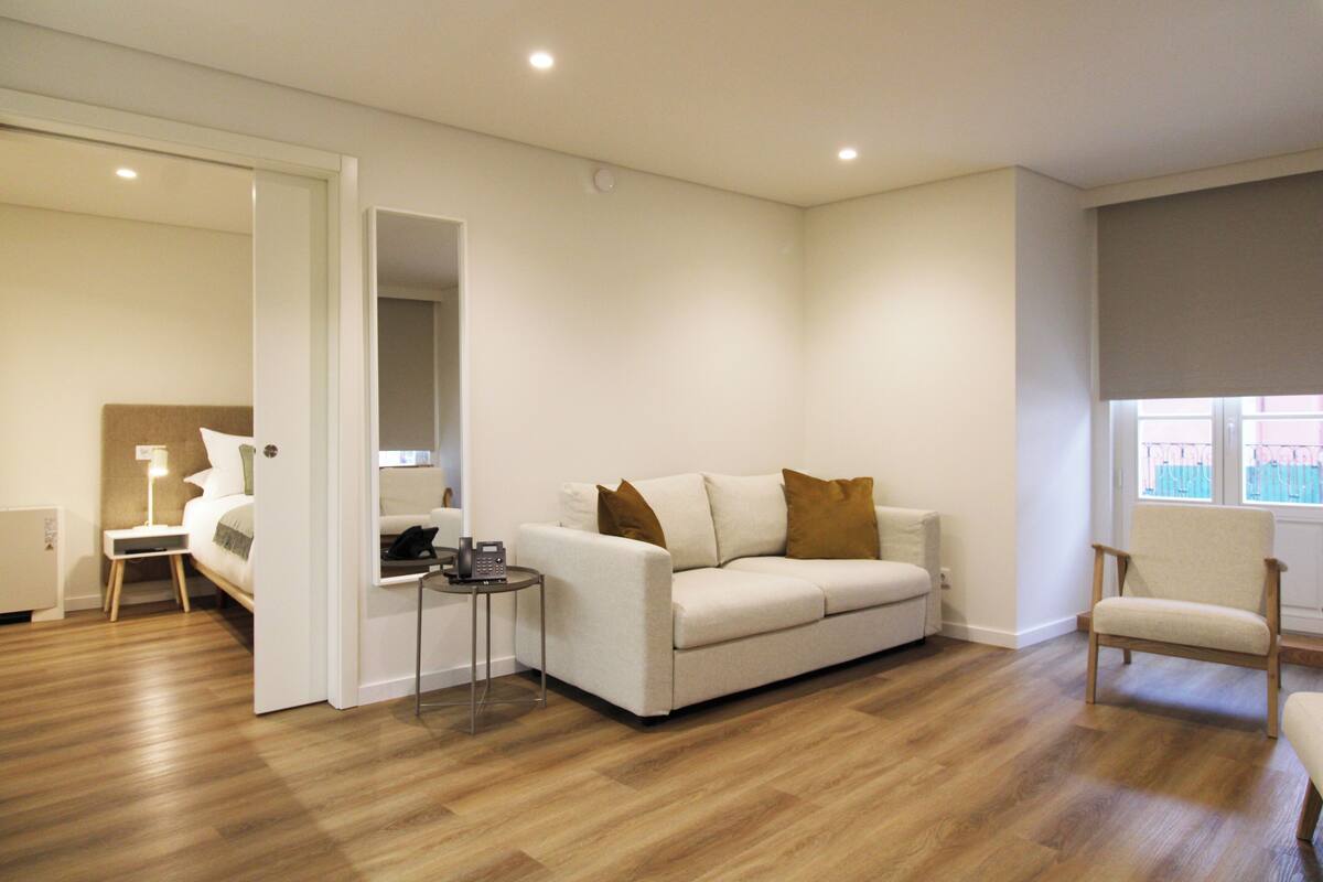 Une chambre d’hôtel moderne avec un canapé blanc, une petite table d’appoint, un plancher en bois et une fenêtre donnant sur un paysage agréable.