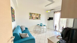 Ein Wohnzimmer mit einem blauen Sofa, einem Esstisch mit Stühlen, einem farbenfrohen Gemälde und einem an der Wand befestigten Fernseher.