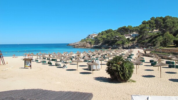Una playa con numerosas tumbonas y sombrillas, una terraza de madera y vistas al mar y a una colina con edificios.