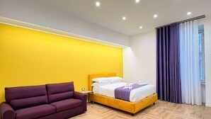A modern bedroom with a wooden herringbone floor, a yellow accent wall, a purple sofa, a bed with white and purple bedding, and a window with purple curtains. - Alla Scoperta di Napoli (Naples)