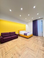 Un dormitorio moderno con suelo de madera en diseño espiga, una pared de acento amarilla, un sofá púrpura, una cama con ropa de cama blanca y morada y una ventana con cortinas moradas.