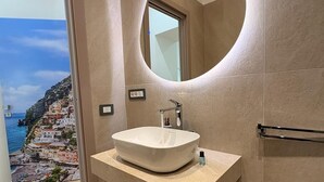 A modern bathroom with a large oval mirror, a white vessel sink, and a view of a coastal town through a large window. - Alla Scoperta di Napoli (Naples)