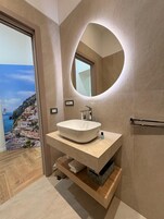 Un baño moderno con un espejo ovalado grande, un lavabo blanco tipo recipiente y vistas a un pueblo costero a través de un ventanal amplio.