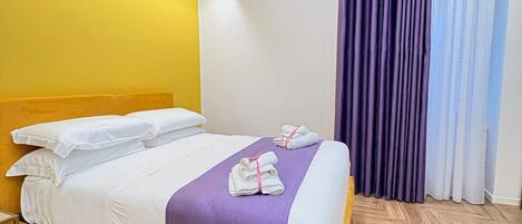 Una habitación de hotel con cama, cabecera, piso de madera y ventana con cortinas.