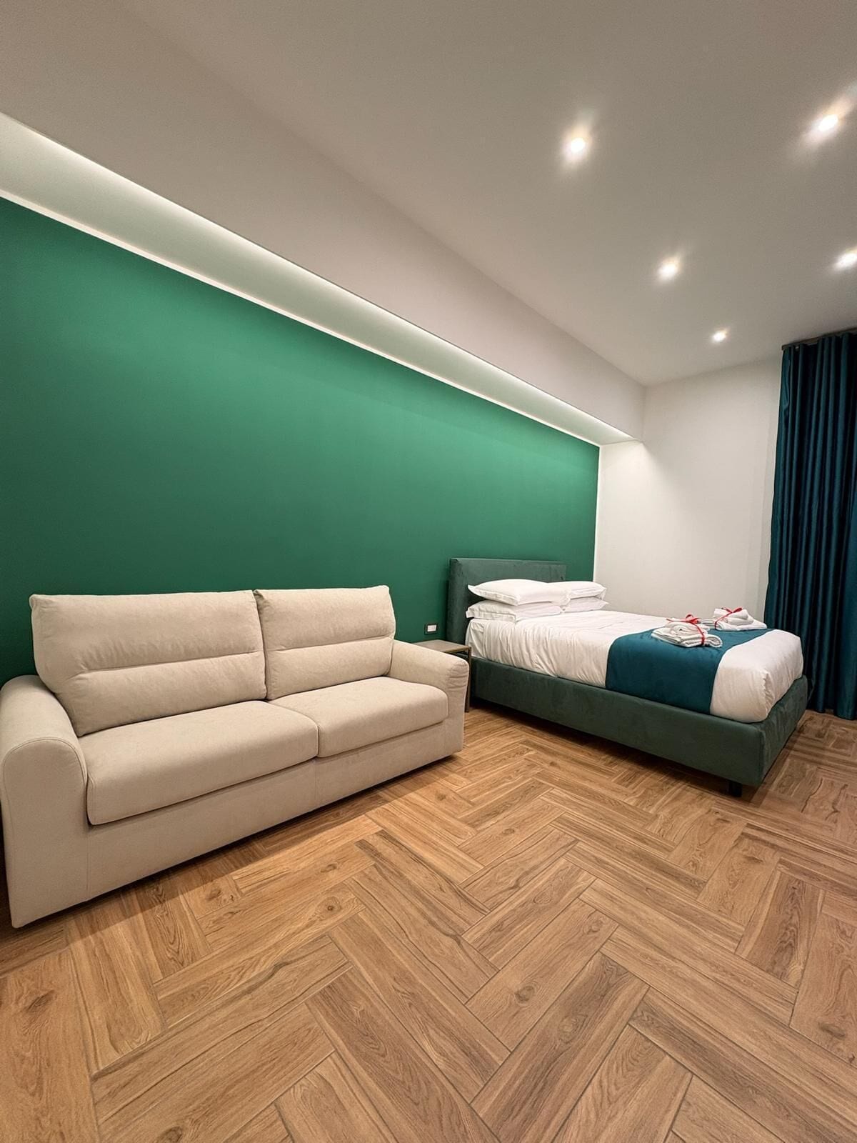 Et moderne hotelværelse med en grøn accentvæg, en beige sofa, en seng med hvide sengetæpper og et trægulv i fiskebensmønster.