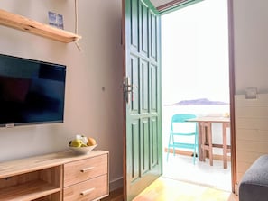Ein Zimmer mit Fernseher, einem Holzschrank, einer Obstschale, einer grünen Tür und Blick aufs Meer.