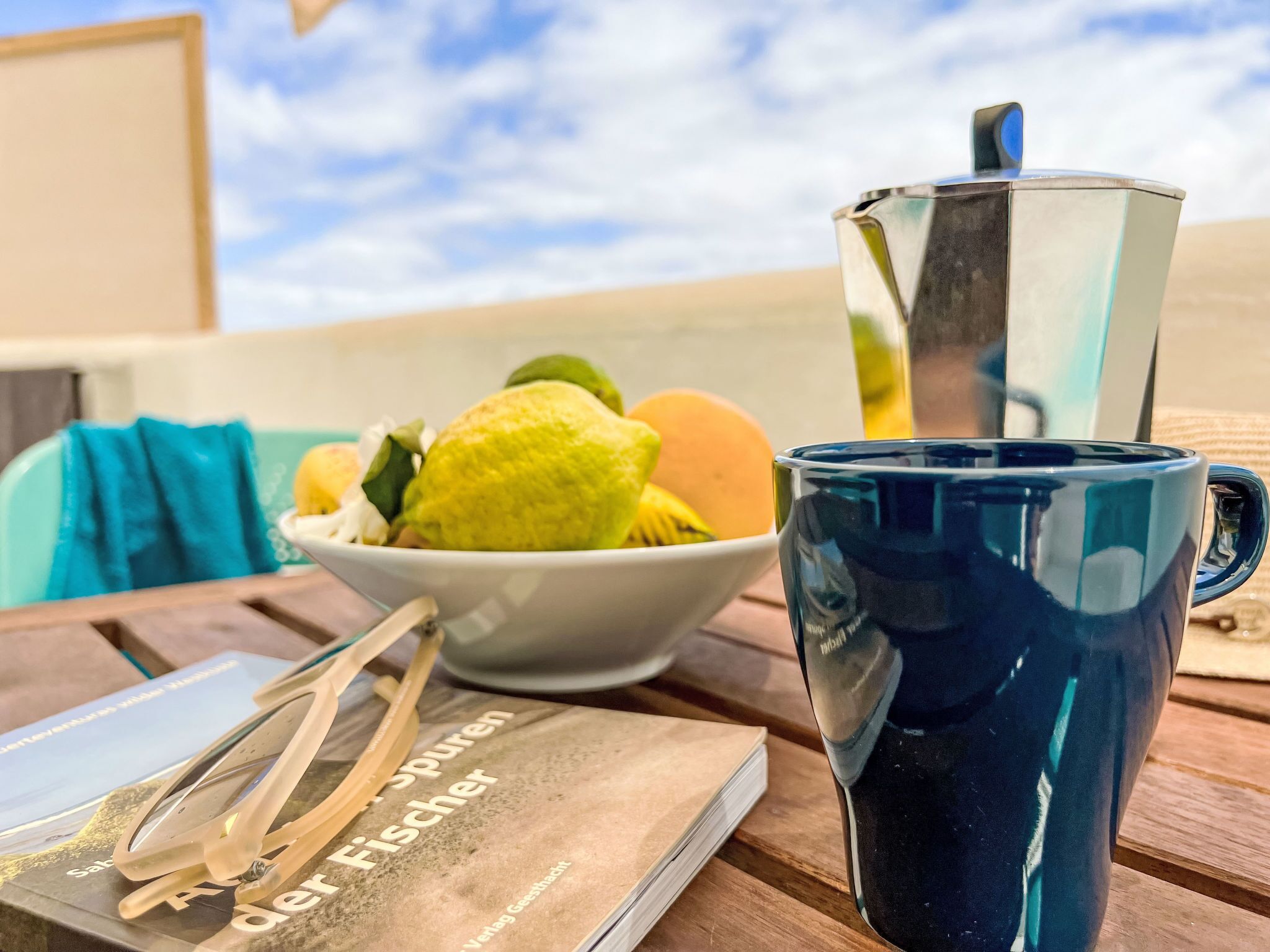 A table with a blue mug, a white bowl with fruit, and a book titled 'Der Fischer'.