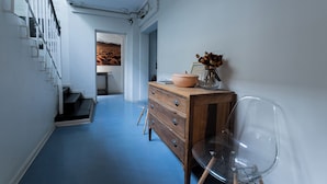 Un pasillo con piso azul, un aparador de madera con cajones, una silla transparente y una escalera con barandillas blancas.