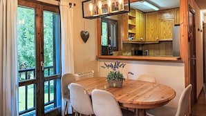 A dining area with a wooden table, white chairs, a potted plant, and a kitchen with wooden cabinets.
