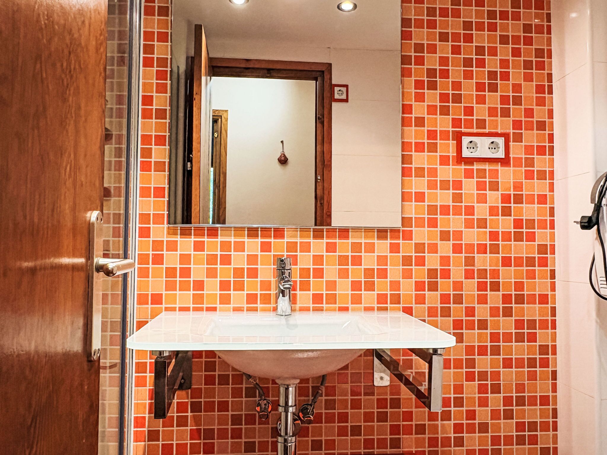 A bathroom with a tiled wall, a sink, a mirror, and a door.