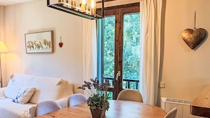 A cozy room with a wooden dining table, white sofa, and a view of greenery through a window.