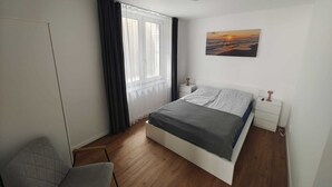 Un dormitorio con cama, una silla, una mesita de noche y un cuadro, con una ventana y cortinas.