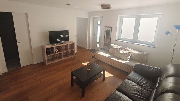 Sala de estar con televisor, piso de madera, sofá de cuero negro, una mesita y una ventana con luz natural.