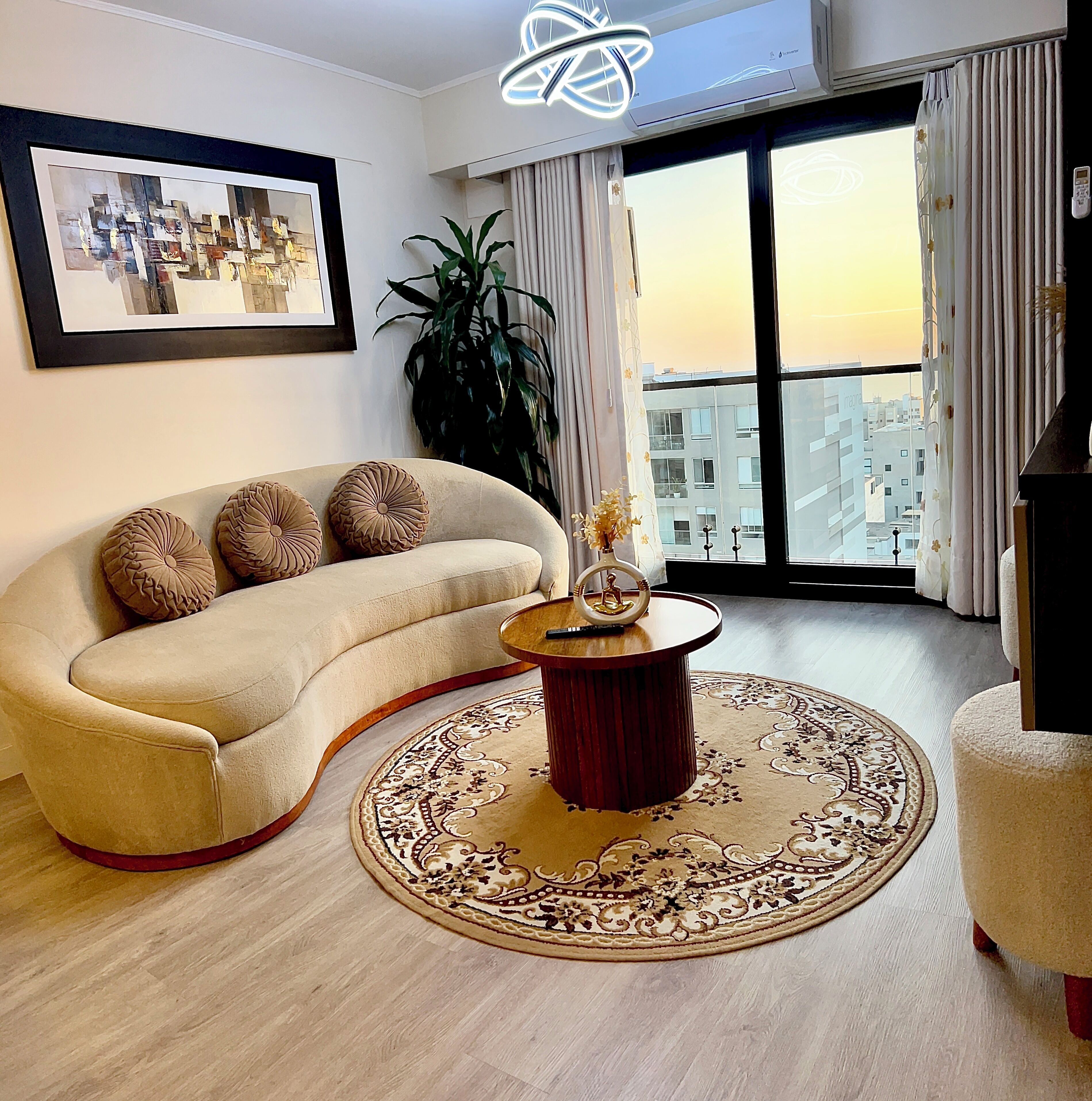 Un salon avec un canapé, une table basse ronde, un tableau encadré au mur et une vue sur les immeubles par une grande fenêtre.