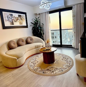 A living room with a sofa, a round coffee table, a framed picture on the wall, and a view of buildings through a large window. - Ocean View | Pool | Gym | A/C | Ravine (Lima)