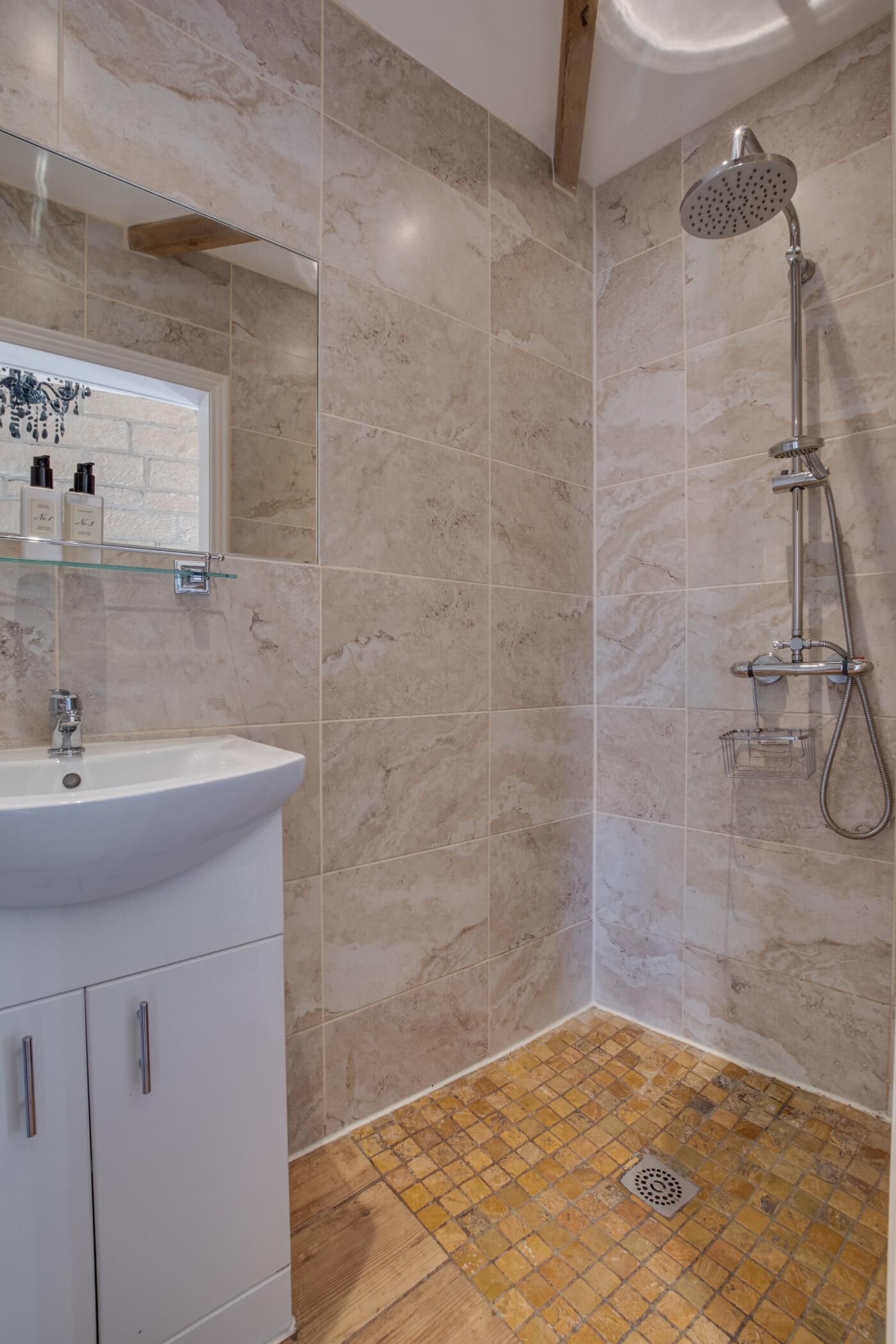 Une salle de bain avec un lavabo blanc, une étagère en verre où sont rangés les articles de toilette, une douche avec un pommeau de douche à main et un sol carrelé avec un drain.