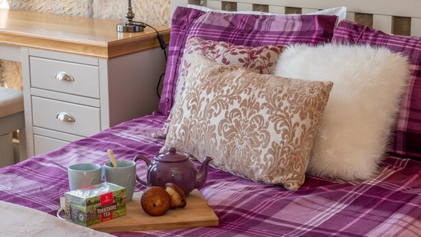 Uma cama bem arrumada com colcha quadriculada em roxo e branco, um travesseiro decorativo, um travesseiro branco fofo, uma pequena bandeja de madeira com um bule, uma xícara, uma caixa de chá Yorkshire e dois muffins.