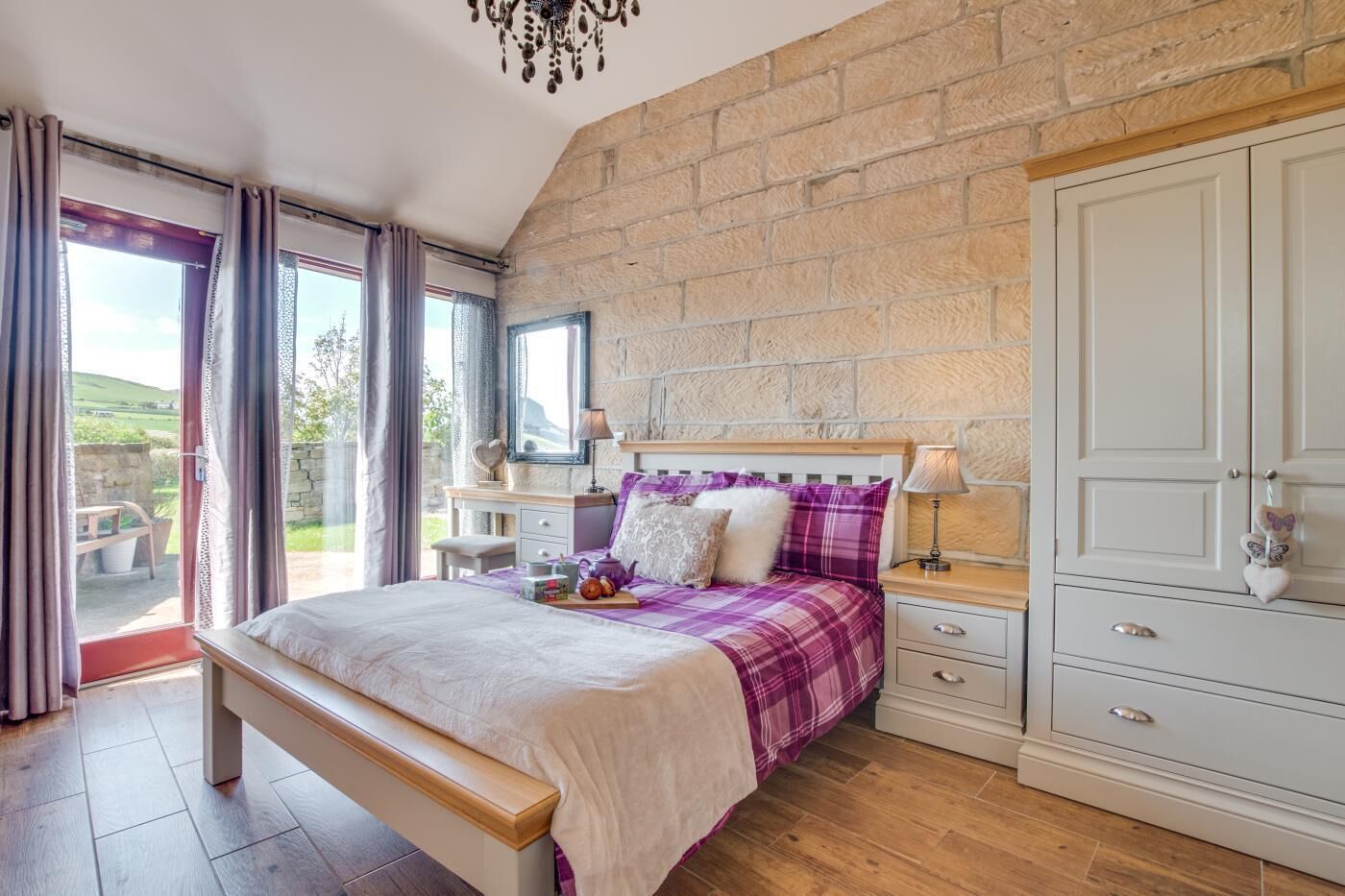 Une chambre avec un mur en pierre, un lit avec une tête de lit en bois, une table de chevet, une commode et une vue sur l’extérieur.