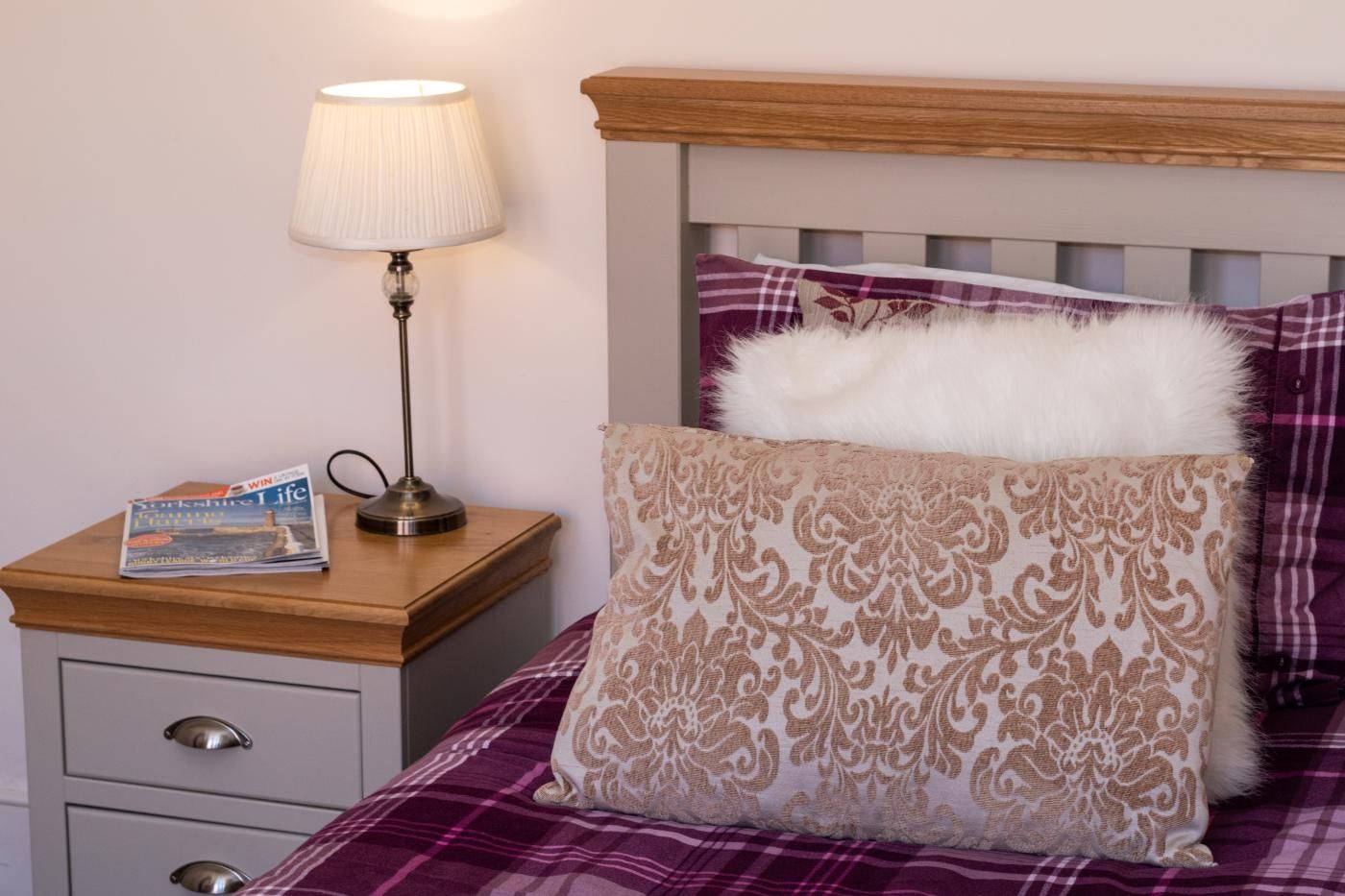 A neatly made bed with a plaid bedspread, a decorative pillow, a bedside lamp, and a magazine on a wooden nightstand.