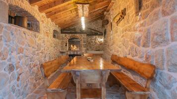 Une pièce aux murs de pierre, avec une table et des bancs en bois, une cheminée et un luminaire suspendu.