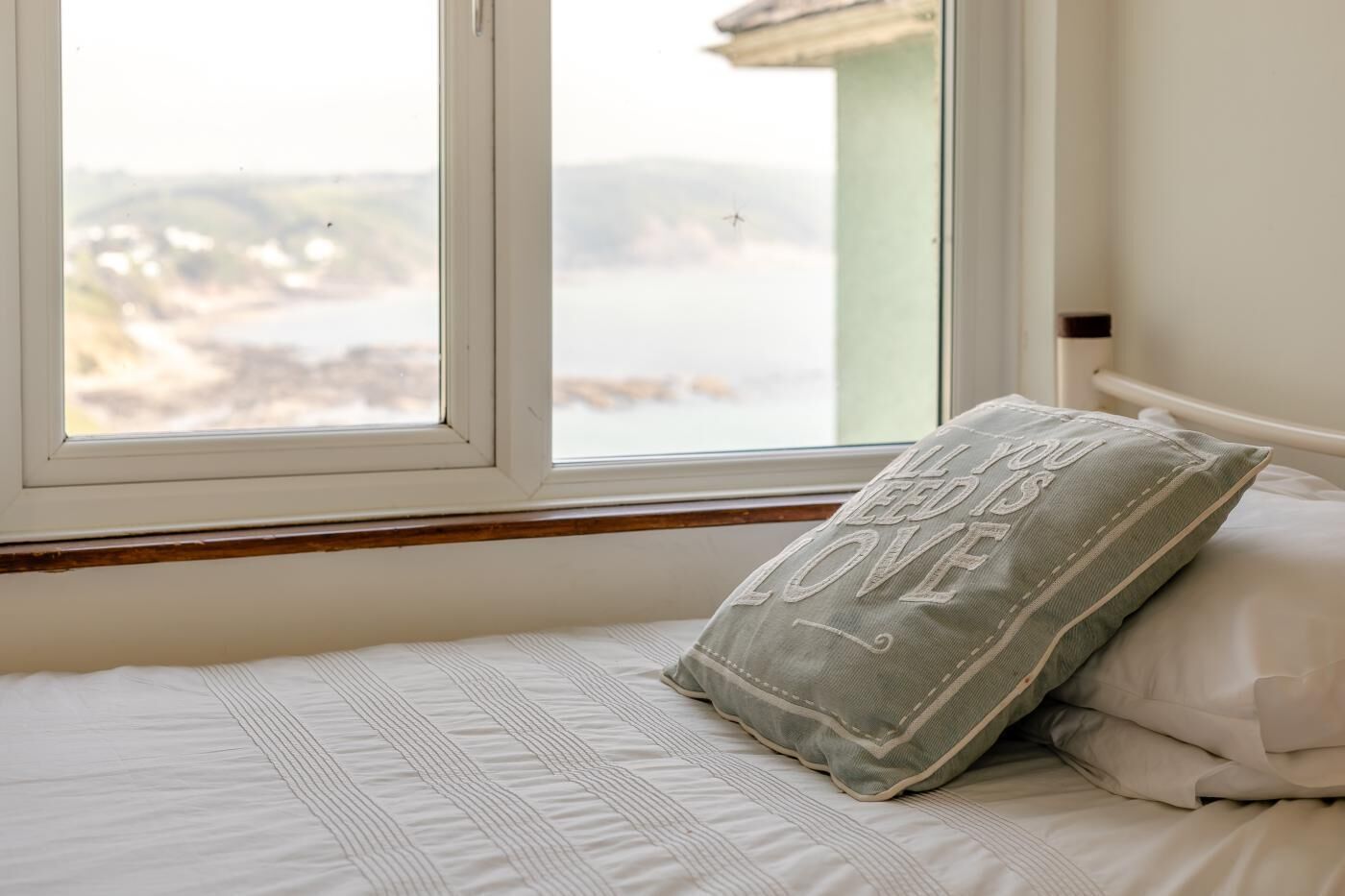 Un lit avec un oreiller sur lequel est brodé le message « TOUT CE DONT VOUS AVEZ BESOIN, C’EST L’AMOUR », et une vue sur la plage par la fenêtre.