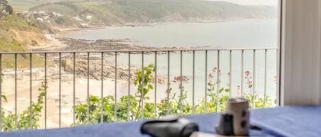 A room with a view of a coastal landscape, a bed with a blue cover, and a window with a metal railing.