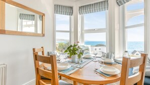 A dining area with a round table, four wooden chairs, a potted plant, and a view of the sea through large windows. - 3 Bed in Looe (oc-l30005) (Looe)