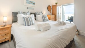 A neatly made bed with white linens and striped pillows, a bedside lamp, a wooden nightstand with drawers, a framed picture on the wall, and a view of the outdoors through a door. - 3 Bed in Looe (oc-l30005) (Looe)