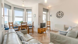 A living room with a sofa, a TV, a dining table with chairs, and a view of the sea. - 3 Bed in Looe (oc-l30005) (Looe)