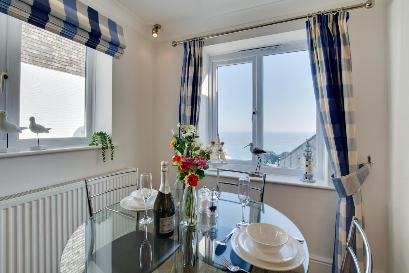 A dining area with a table set for two, a bottle of wine, and a vase of flowers. There are two windows with curtains, one of which has a view of the sea.