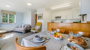 A dining area with a table set for two, a sofa, a recliner, and a kitchen in the background. - 2 Bed in Looe (oc-l00055) (Trelawne)