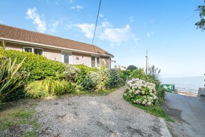 A house with a gravel driveway, lush greenery, and a view of the sea. - 2 Bed in Looe (oc-l00055) (Trelawne)