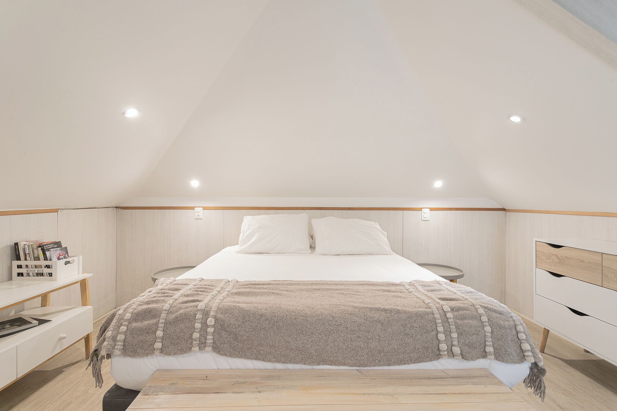 Un dormitorio con techo inclinado, cama con edredón blanco y manta color beige, mesitas de noche de madera y una lámpara de noche pequeña.