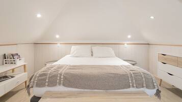 Un dormitorio con techo inclinado, cama con edredón blanco y manta color beige, mesitas de noche de madera y una lámpara de noche pequeña.