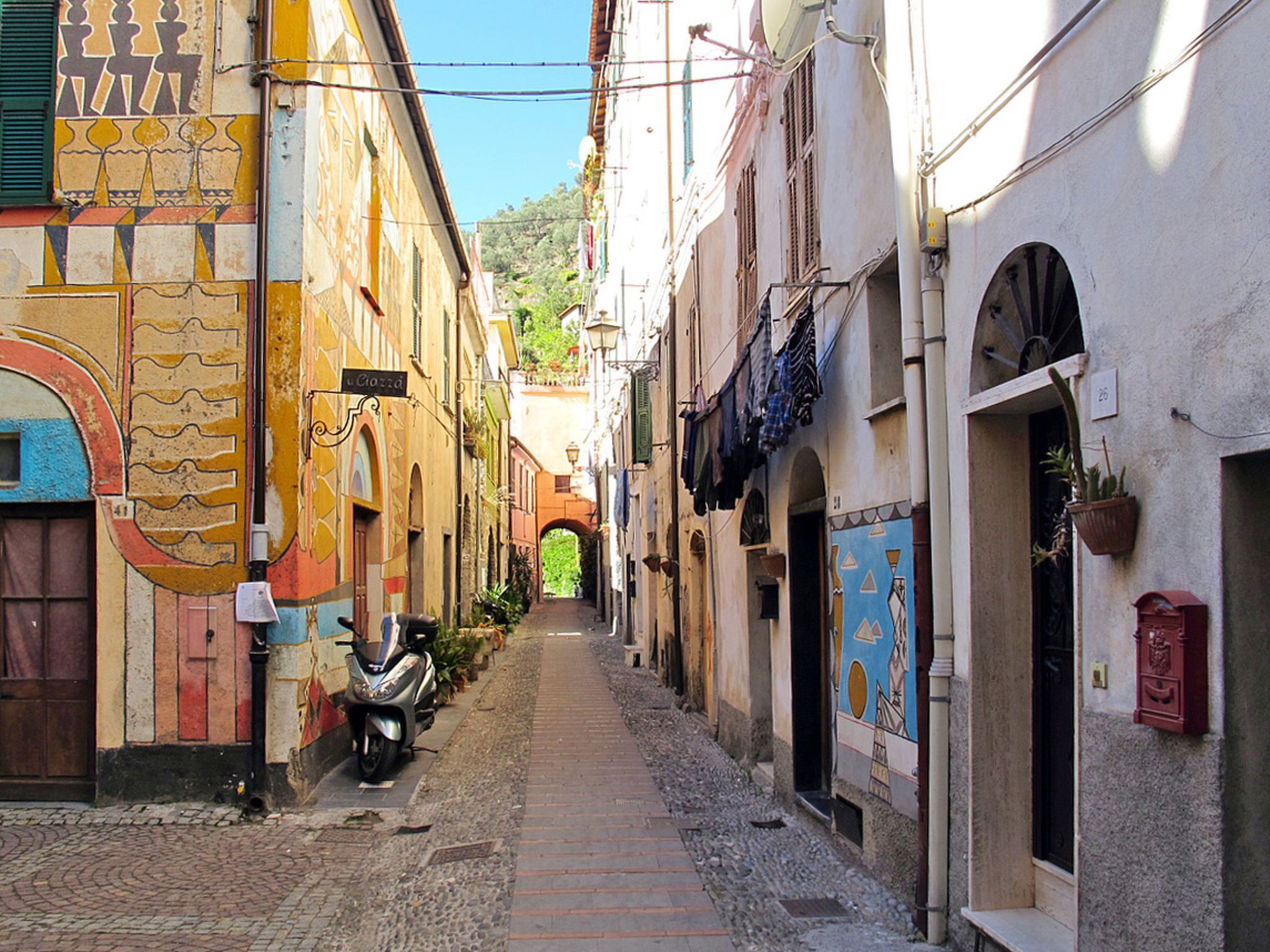 A narrow cobblestone street lined with colorful buildings, a scooter parked on the side, and an archway in the distance.