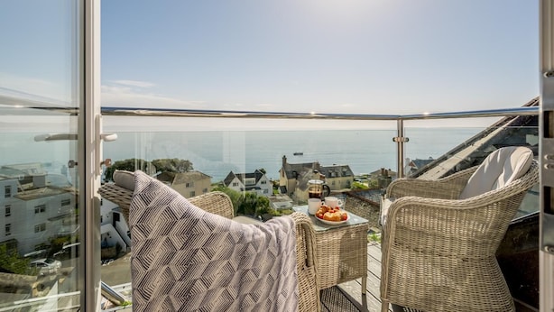 A balcony with wicker furniture, a table with a plate of pastries, and a view of the sea and buildings. - 2 Bed in Looe (oc-l30011) (Looe)