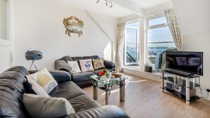 A modern living room with a leather sofa, a glass coffee table, a flat-screen TV, and a large window offering a view of the sea. - 2 Bed in Looe (oc-l30011) (Looe)