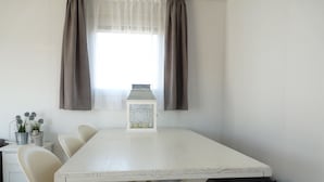 Een strak ingerichte kamer met een witte tafel, beige stoelen, een witte kast met een lantaarn en een raam met gordijnen.