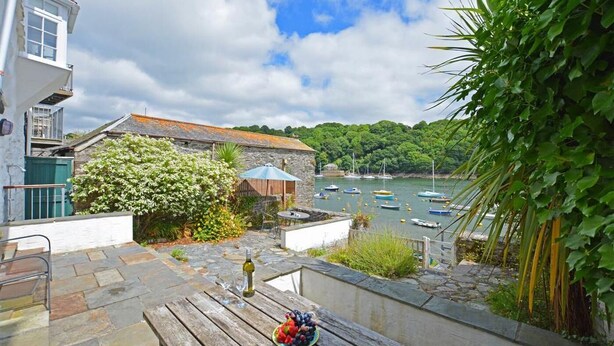 Um alpendre de pedra com uma mesa posta para refeição, com vista para um porto cheio de barcos e vegetação.