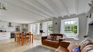 Uma sala de estar espaçosa com uma mesa de jantar e cadeiras, um sofá de couro marrom com almofadas e grandes janelas com vista para o verde.