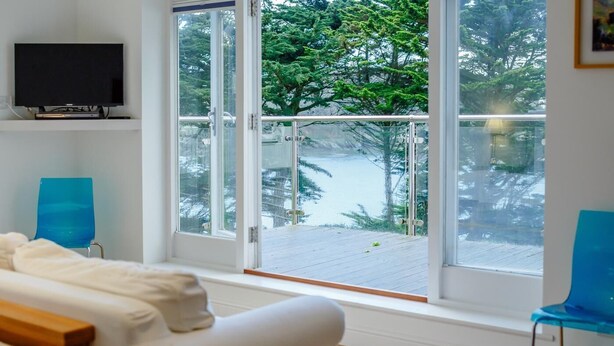 A room with a large window overlooking a snowy deck and trees, a white sofa, and two blue chairs. - 5 Bed in Polruan (oc-ccmead) (Fowey)