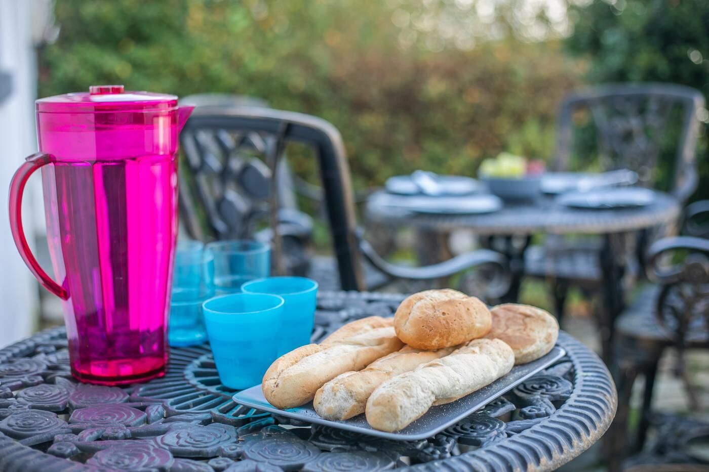 A table with a plate of bread rolls, a pink pitcher, and blue cups.