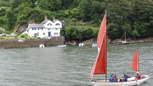 Un velero con pasajeros en un río tranquilo, una casa blanca con puerta azul en la orilla, y una colina boscosa al fondo.