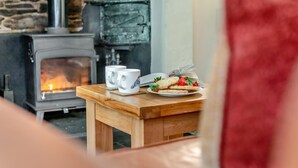 Un cuarto acogedor con una estufa de leña, una mesa de madera con un plato de comida y dos tazas, y una cortina roja visible en parte.