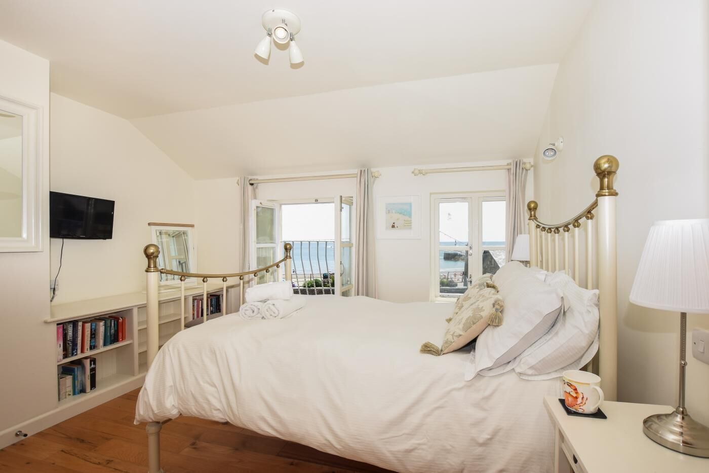 A bedroom with a large bed, a bedside table with a lamp, a bookshelf, and a view of the sea through a window.