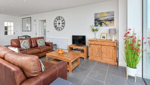 A modern living room with a brown leather sofa, a wooden coffee table with a fruit bowl, a television, and a wooden cabinet. - 5 Bed in Pwllheli (oc-dolwen) (Llanbedrog)