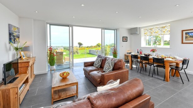 A modern living room with a leather sofa, a wooden coffee table, and a dining area with a table and chairs. - 5 Bed in Pwllheli (oc-dolwen) (Llanbedrog)